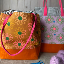 Load image into Gallery viewer, Two fabric tote bags with a floral daisy pattern are shown on a tabletop. The front bag is mustard yellow with pink and peach flowers, green centers, and a bright pink handle and orange base.