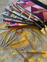 Load image into Gallery viewer, Collection of knitting needles and colorful case on a textured fabric background