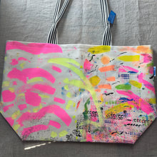 Load image into Gallery viewer, A canvas tote bag with black and white striped handles, featuring a vibrant abstract paint splatter pattern in pink, yellow, green, and orange on a white background, laid flat on a gray fabric surface.

